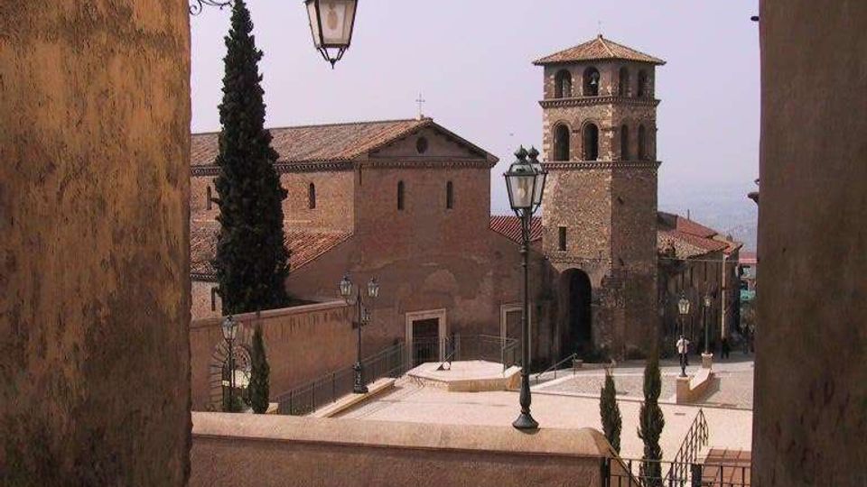 Veduta di una piazza in un borgo storico con pavimentazione in pietra, lampioni in ferro battuto e muretti, dominata da una chiesa in mattoni con campanile a torre e tetti in coppi. La scena è incorniciata da edifici alti ai lati e da alcuni cipressi lungo il percorso in discesa.