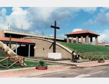 Area esterna di un complesso commemorativo con una grande croce in legno su basamento, vialetti pavimentati e aiuole fiorite. Sullo sfondo si vedono edifici in mattoni con tetti in tegole e un pendio erboso con staccionate in legno.