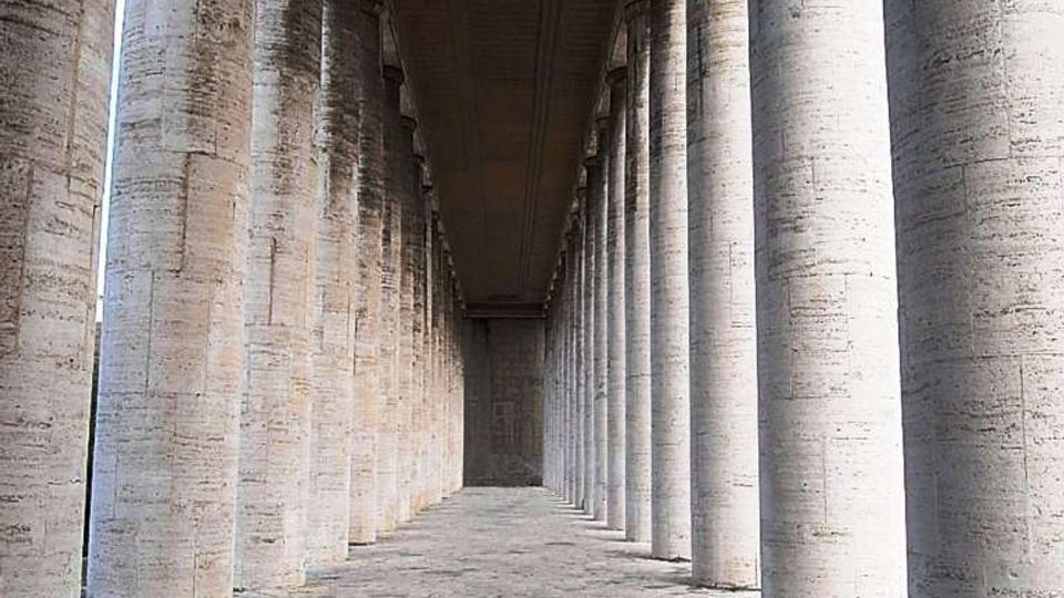 Lungo porticato monumentale con alte colonne cilindriche in pietra disposte in file parallele, che creano una prospettiva verso il fondo. Pavimentazione in lastre di pietra irregolari e soffitto piano in cemento sopra il corridoio.
