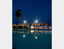 Piscina esterna di un hotel al crepuscolo, con acqua calma che riflette le luci dell’edificio moderno a due piani. A bordo piscina si vedono lettini e ombrelloni chiusi, una palma e grandi vetrate illuminate sul lato della struttura.