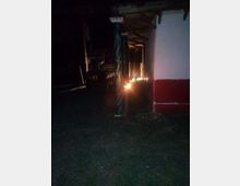Veranda esterna di notte con tetto in legno sorretto da pali, affiancata a un edificio bianco con fascia rossa alla base. In primo piano c’è un prato e sotto la tettoia si vedono arredi e una piccola fonte di luce vicino a uno dei pilastri.