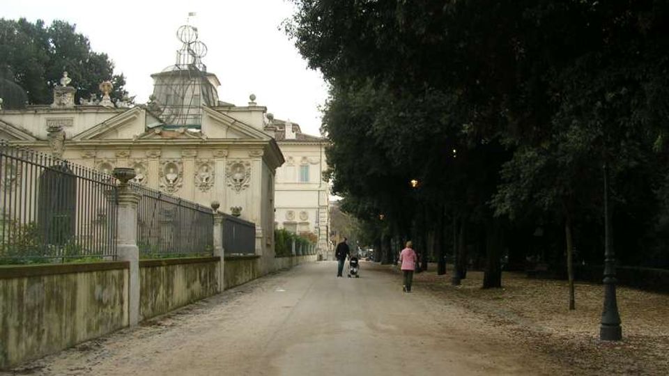 Viale pedonale in un parco, con ampia strada sterrata fiancheggiata da alti alberi e lampioni sul lato destro. A sinistra si vede un edificio storico decorato con facciata in stile classico e recinzione in ferro sopra un muretto.