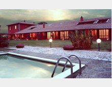 Piscina all’aperto con bordo in cemento e scaletta metallica in primo piano, affacciata su un vialetto ghiaioso e un prato con lampioni sferici. Sullo sfondo si vede un edificio basso con tetto in tegole e grandi vetrate illuminate, in un contesto rurale al tramonto.