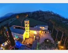 Veduta aerea al crepuscolo di un complesso rurale in pietra con campanile di una piccola chiesa illuminata da luci calde, tetti in coppi e cortile interno. Intorno si vedono colline boscose e campi, con alti pini e un viale che conduce alla struttura.