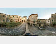 Piazza in un borgo storico con pavimentazione in pietra e gradinate curve che scendono verso un’area centrale. Intorno si vedono edifici antichi in muratura intonacata e pietra, con finestre e balconi, e passaggi ad arco tra le case.