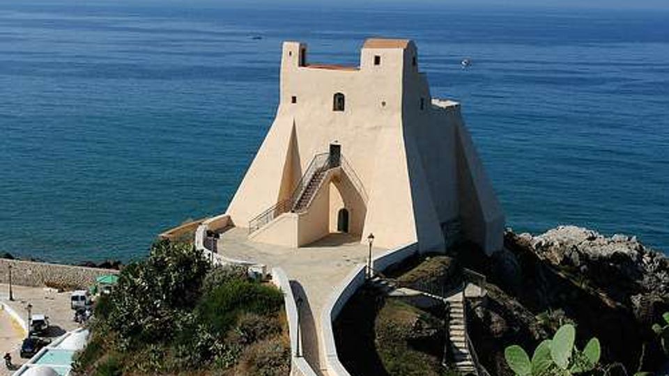 Fortezza costiera in pietra chiara con mura inclinate e piccola torre, arroccata su uno sperone roccioso affacciato sul mare. Una passerella e una scalinata conducono all’ingresso, con il litorale e l’orizzonte marino sullo sfondo.