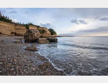 Spiaggia di ciottoli lungo una costa con falesie sabbiose e vegetazione bassa, con grandi rocce affioranti vicino alla riva. Il mare calmo si estende verso un molo lontano con alcuni pali di illuminazione sotto un cielo nuvoloso.
