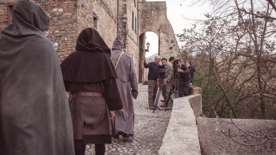 Stradina acciottolata lungo un edificio in pietra e mattoni con un arco e una lanterna, affiancata da un parapetto che si affaccia su alberi spogli in un paesaggio collinare. Sullo sfondo si vedono attrezzature cinematografiche posizionate sul percorso.