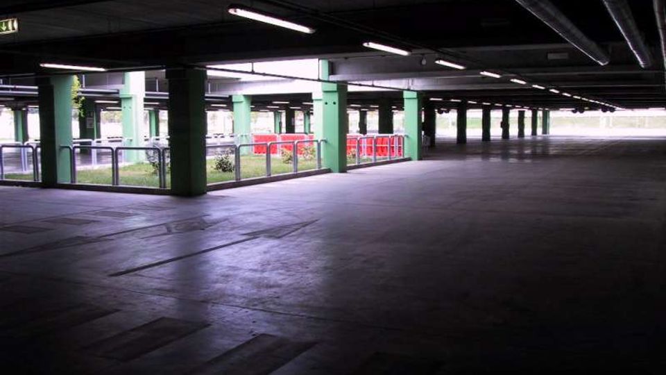 Ampio parcheggio coperto con pavimento in cemento, soffitto basso con tubazioni a vista e lunghe luci fluorescenti. Pilastri verdi scandiscono lo spazio, con ringhiere metalliche e alcune barriere rosse vicino alle aperture verso l’esterno.