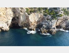 Scogliera rocciosa a picco sul mare con una grande grotta naturale alla base e speroni di roccia frastagliati lambiti dalle onde. Acqua blu scura in primo piano e vegetazione rada sulla sommità della parete calcarea.