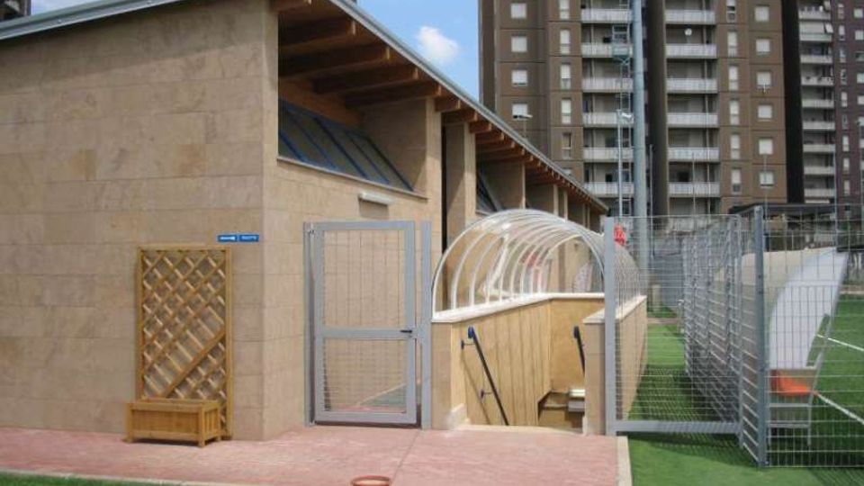 Edificio basso in muratura chiara con tetto a falda e travi a vista, affiancato da un vialetto in mattoni che conduce a una rampa di scale coperta da una pensilina trasparente. A destra si vede un’area sportiva con erba sintetica recintata da alte reti metalliche, con sullo sfondo palazzi residenziali.