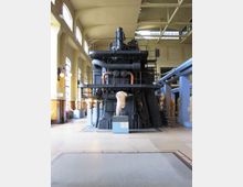 Interno di un museo tecnico in una grande sala industriale con alte finestre ad arco e soffitto a capriate. Al centro si trova un enorme macchinario metallico scuro con tubazioni e passerelle, circondato da pannelli espositivi e basamenti.