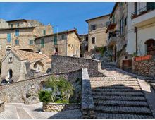 Scalinata in pietra che sale tra case in muratura con facciate chiare, persiane e balconi, in un borgo storico collinare. Muri e pavimentazioni in pietra delimitano piccoli terrazzamenti con vegetazione sotto un cielo sereno.