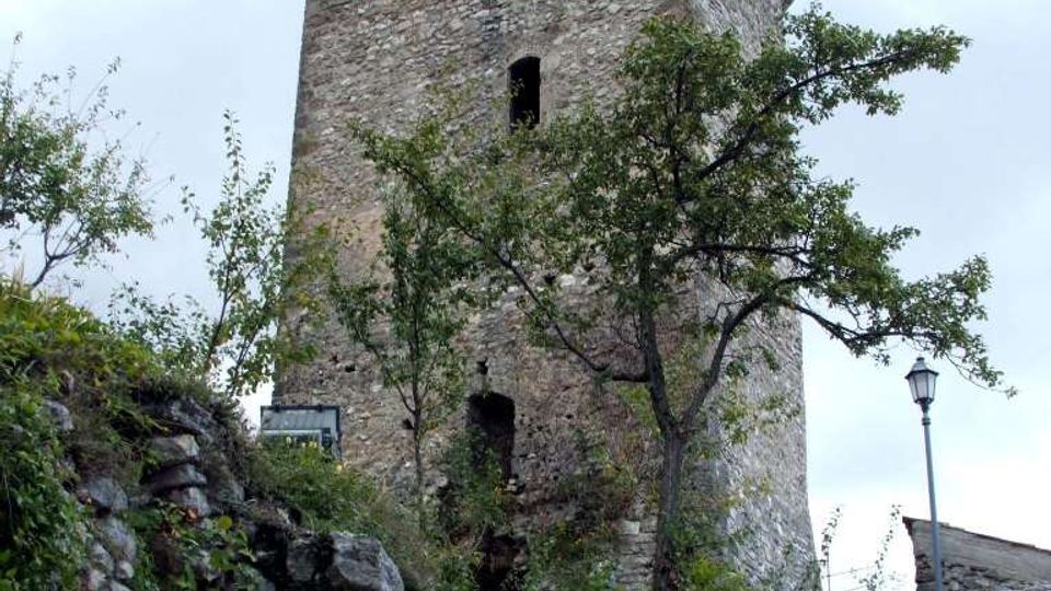Torre medievale in pietra con merlature sulla sommità e piccole aperture, vista dal basso. In primo piano una scalinata di pietra tra rocce e vegetazione conduce verso la torre, con un lampione e alberi ai lati.
