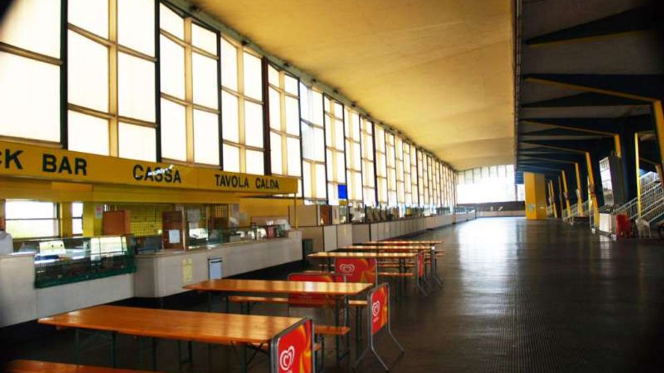 Ampio spazio interno tipo mensa o area ristoro in un edificio moderno, con grandi finestre a tutta altezza lungo un lato e soffitto alto. In primo piano ci sono tavoli e panche in legno disposti in file, mentre sul lato sinistro si vedono i banconi del bar e della tavola calda.