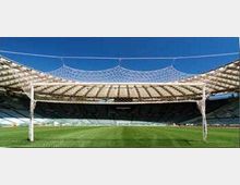 Interno di uno stadio all’aperto visto dal campo, con ampia area di prato verde e tribune semicircolari sullo sfondo. Copertura leggera con struttura reticolare e pilastri bianchi che sorreggono una rete sopra la zona centrale.