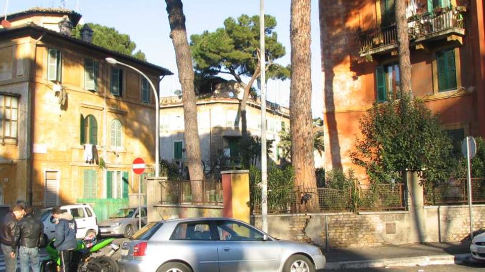 Strada urbana con auto parcheggiate davanti a palazzi residenziali dai toni ocra e rosso, con persiane verdi e balconi in ferro. Alti pini marittimi e un muro con recinzione delimitano il marciapiede, con segnaletica stradale visibile.