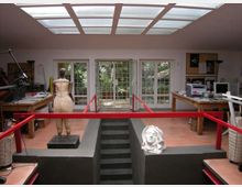 Interno di un laboratorio o studio artistico con lucernario a griglia sul soffitto, porte-finestre vetrate sul fondo e tavoli da lavoro ai lati. Al centro c’è una piattaforma rialzata con scala e parapetti rossi, con un manichino e una scultura di testa appoggiata.