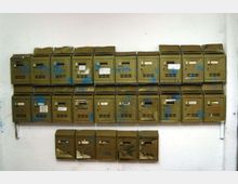 Fila di cassette postali metalliche verde oliva fissate a una parete bianca, disposte su tre livelli; molte hanno etichette, fessure per la posta e segni di usura o vernice.