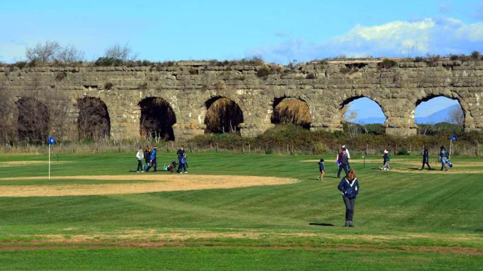 Campo da golf con ampie distese di erba e una buca segnalata da bandierina, con sullo sfondo un lungo acquedotto in pietra con grandi arcate. Terreno leggermente ondulato in ambiente aperto, con vegetazione bassa e cielo azzurro.