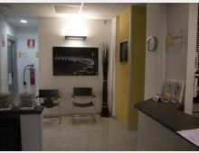 Piccola sala d’attesa interna con pavimento piastrellato e controsoffitto, due sedie metalliche, quadro a parete e illuminazione a plafoniera. A destra si vede un bancone reception, un pannello divisorio giallo con schermo integrato e un orologio a parete, con estintore e cartelli di sicurezza sul lato sinistro.