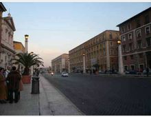 Ampio viale urbano al tramonto con carreggiata in asfalto scuro e marciapiede in pietra, fiancheggiato da lampioni e grandi palme in vaso. Ai lati si trovano alti edifici storici in muratura con facciate uniformi e finestre regolari, con alcune auto in transito lungo la strada.
