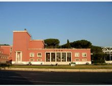 Edificio basso in stile razionalista con facciata rosa, ingresso centrale con grandi vetrate e gradinata, affiancato da un volume più alto sul lato sinistro. Davanti c’è una strada ampia con prato e alcuni veicoli, sullo sfondo alberi e un ponte metallico sulla destra.
