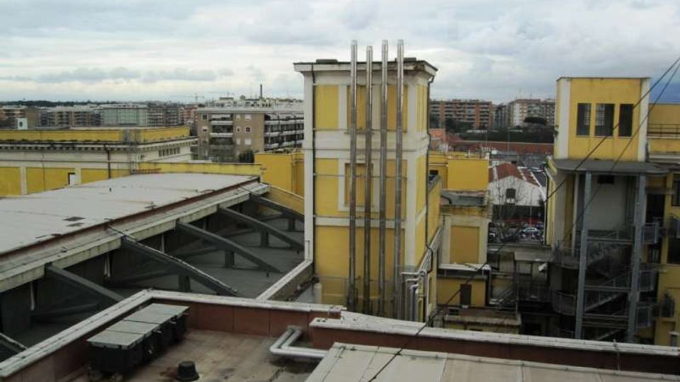 Vista su un complesso di edifici industriali gialli con tetti piani, passerelle coperte e condotte verticali in metallo su una torre centrale. Sullo sfondo si vedono palazzi residenziali e un cielo nuvoloso.