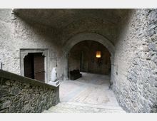 Cortile interno in pietra con soffitto a volta e grande arco che conduce a un corridoio coperto, illuminato da una piccola finestra. Sulla sinistra si vede una porta in legno incorniciata da pietra e una breve scalinata con muretto in muratura.