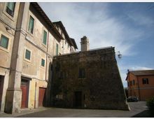 Cortile urbano con edifici in muratura intonacata e pietra: a sinistra una palazzina a più piani con finestre verdi e portoni al piano terra, al centro un edificio più basso in pietra scura con piccolo comignolo. Sullo sfondo si vede una strada con una casa moderna e un lampione sotto un cielo parzialmente nuvoloso.