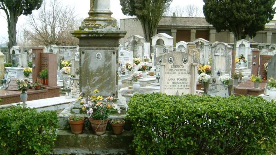 Cimitero con numerose tombe e lapidi in pietra disposte in file, alcune decorate con fiori e vasi. In primo piano si vede un monumento funerario con base in pietra accanto a siepi basse, con alberi alti sullo sfondo.