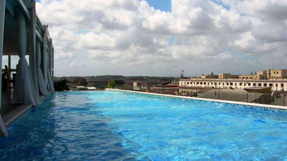Piscina a sfioro su una terrazza panoramica, con acqua azzurra e bordo bianco affacciato su un paesaggio urbano di edifici bassi. A sinistra si vede una struttura coperta con tende leggere, sotto un cielo ampio con nuvole.