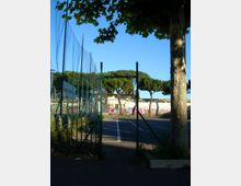 Ingresso a un campo sportivo recintato con rete metallica e pali alti, con un vialetto asfaltato che conduce a un’area di gioco segnata da linee bianche. Sullo sfondo si vedono pini e un edificio basso con graffiti, sotto un cielo sereno.