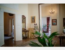 Interno di una casa in stile classico con pavimento in parquet e porte bianche alte che collegano più stanze. Sono visibili un lampadario a bracci, quadri alle pareti, un piccolo tavolo con specchiera, tappeti e una finestra con tende a righe rosse e verdi accanto a piante in vaso.