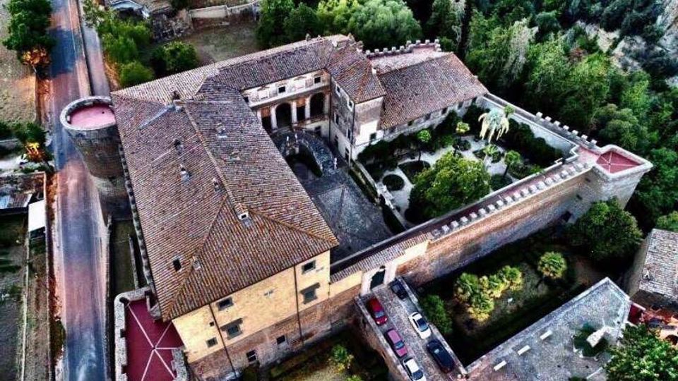Veduta aerea di un grande complesso in muratura con tetti in coppi, organizzato attorno a un cortile interno e circondato da mura merlate con torri agli angoli. All’interno delle mura si vede un giardino alberato e, all’esterno, una strada che corre lungo il lato sinistro dell’edificio.