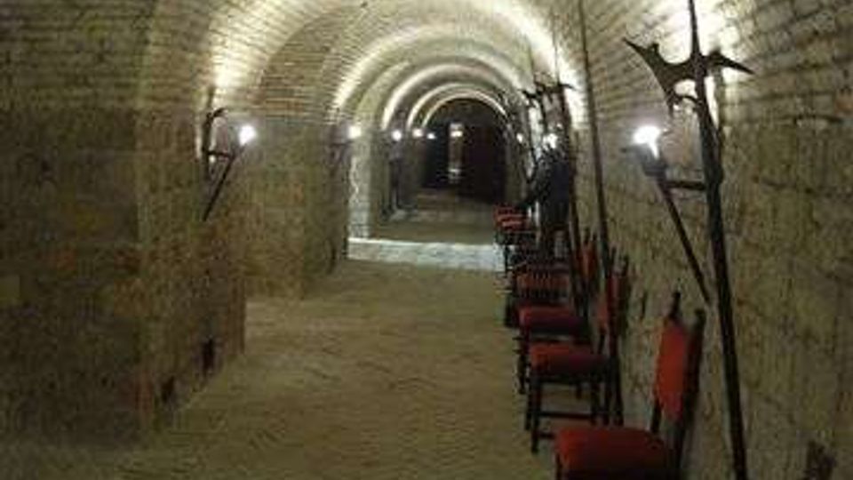 Corridoio sotterraneo in pietra con soffitto a volta a botte, illuminato da lampade a parete. Lungo una parete sono disposte sedie e armi antiche esposte, con il passaggio che si estende fino a un’apertura in fondo.