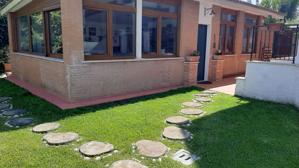 Esterno di una casa a un piano in mattoni con tetto in tegole e grandi finestre con infissi in legno. Davanti c’è un prato curato con un percorso di lastre circolari in pietra e un piccolo ingresso con aiuole e ringhiera metallica.