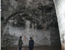 Interno di un ambiente sotterraneo in cemento, simile a un bunker, con soffitto a volta e pareti grezze macchiate e scrostate. Sulla parete di fondo si notano una nicchia ad arco, fori e piccoli graffiti, con illuminazione fioca e ombre marcate.