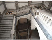 Interno di un vano scala monumentale in marmo con rampe sovrapposte, balaustre scolpite e ampi archi decorati. Pareti rivestite in pietra e pavimenti a motivi geometrici con corrimani in metallo lungo i gradini.