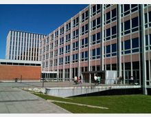 Complesso di edifici moderni per uffici o università con facciate a griglia di grandi finestre e pannelli in laterizio, affacciato su un’ampia area pavimentata con prato. In primo piano si vede una rampa o accesso ribassato con ringhiere metalliche sotto un cielo sereno.