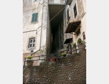 Vicolo stretto tra edifici in muratura e intonaco invecchiato, con una scalinata ripida che sale in un passaggio coperto tra le case. In primo piano un muretto in pietra con ringhiera e vasi di piante, e finestre con persiane e grate sulle facciate.