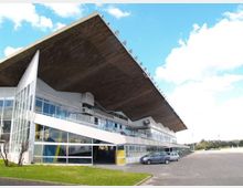 Edificio moderno a più livelli con ampie vetrate e una grande copertura sporgente inclinata, affacciato su un’ampia area asfaltata tipo parcheggio. In primo piano si vedono una striscia di prato e alcune auto, con cielo azzurro sullo sfondo.