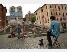 Piazza pavimentata in pietra con ampia scalinata monumentale che sale verso una chiesa con due campanili in restauro e teli protettivi. Ai lati si vedono palazzi storici con finestre e persiane, una fontana con vasca e balaustre in pietra.