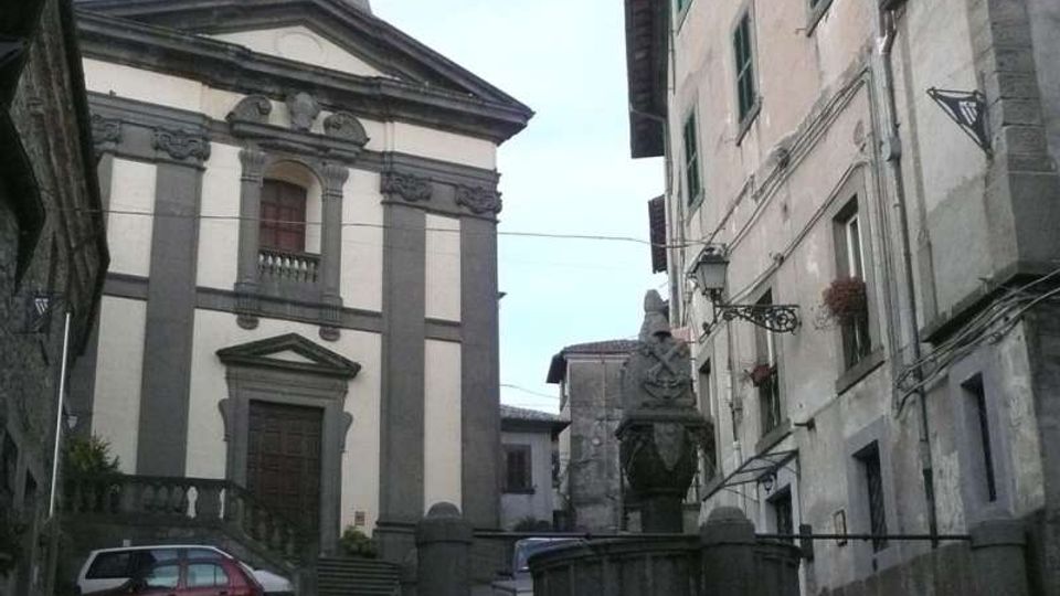 Piazza di un borgo con una chiesa in facciata classica a due colori e portale centrale, affiancata da edifici in pietra con finestre e lampioni. In primo piano si vede una fontana monumentale in pietra con vasca e decorazioni scolpite.