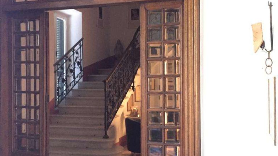 Grande portale interno ad arco in legno con riquadri di vetro, che introduce a un atrio con ampia scala in pietra e ringhiera in ferro battuto. In primo piano si vede un pavimento a mosaico decorato e, a lato, un grande vaso in ceramica contro una parete chiara.