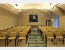Sala conferenze con soffitto a volta e pareti in intonaco chiaro, illuminata da faretti su binari. File di sedie beige disposte ai lati di un corridoio centrale con pavimento in vetro, con un tavolo e un grande pannello appeso sulla parete di fondo.