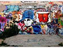 Muro esterno in cemento ricoperto da graffiti e murales multicolori, con un grande disegno centrale di un volto stilizzato con cappellino blu e numerose scritte sovrapposte. Davanti al muro c’è un’area pavimentata in cemento con segni e vernice, ai margini qualche ciuffo di vegetazione.