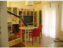 Soggiorno moderno open space con angolo cottura a sinistra, tavolo rotondo bianco con sedie rosse e libreria a parete con TV. A destra una grande portafinestra con tende bianche illumina il pavimento chiaro; in primo piano si vede la ringhiera di una scala interna.