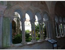 Chiostro in pietra con galleria ad archi a sesto acuto e colonne scolpite, affacciata su un cortile interno con prato e vegetazione. In primo piano si vede un pannello informativo e, sullo sfondo, le mura dell’edificio storico.