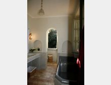 Bagno con pareti chiare e pavimento in pietra, con lavabo in muratura a sinistra e grande vasca scura a destra. Al centro una finestra ad arco aperta sul verde esterno, con nicchie ad arco e lampada a sospensione sul soffitto.